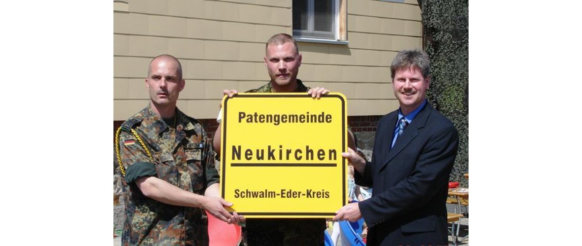 Bürgermeister Olbrich hält mit zwei Soldaten ein Ortsschild ähnliches gelbes Schild mit der Aufschrift "Patengemeinde Neukirchen Schwalm Eder Kreis"