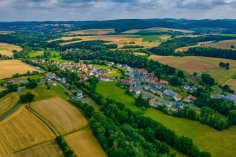 Luftaufnahme Nausis Luftaufnahme des Stadtteils Nausis im Sommer. Im Vordergrund sieht man einen Wald, links davon einige Felder, rechts davon eine Wiese, im Hintergrund eine hügelige Landschaft unter einem bedeckten Himmel.