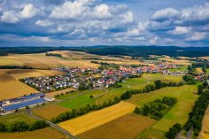 Luftaufnahme Riebelsdorf Luftaufnahme des Stadtteils Riebelsdorf im Sommer. Im Vordergrund einige Felder und Bäume, im Hintergrund eine hügelige Landschaft unter einem Himmel mit großen Wolken.
