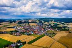 Luftaufnahme Seigertshausen Luftaufnahme des Stadtteils Riebelsdorf im Sommer. Im Vordergrund sieht man einige abgeerntete Felder, im Hintergrund eine Hügellandschaft unter bedecktem Himmel.