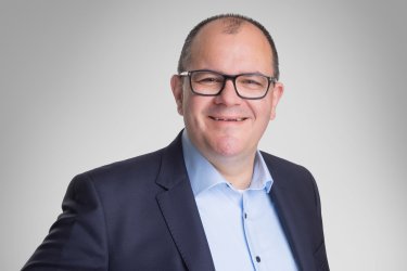 Bild von Bürgermeister Marian Knauf im dunkelblauem Anzug und hellblauen Hemd in der Mitte des Bildes. Der Hintergrund ist hellgrau.