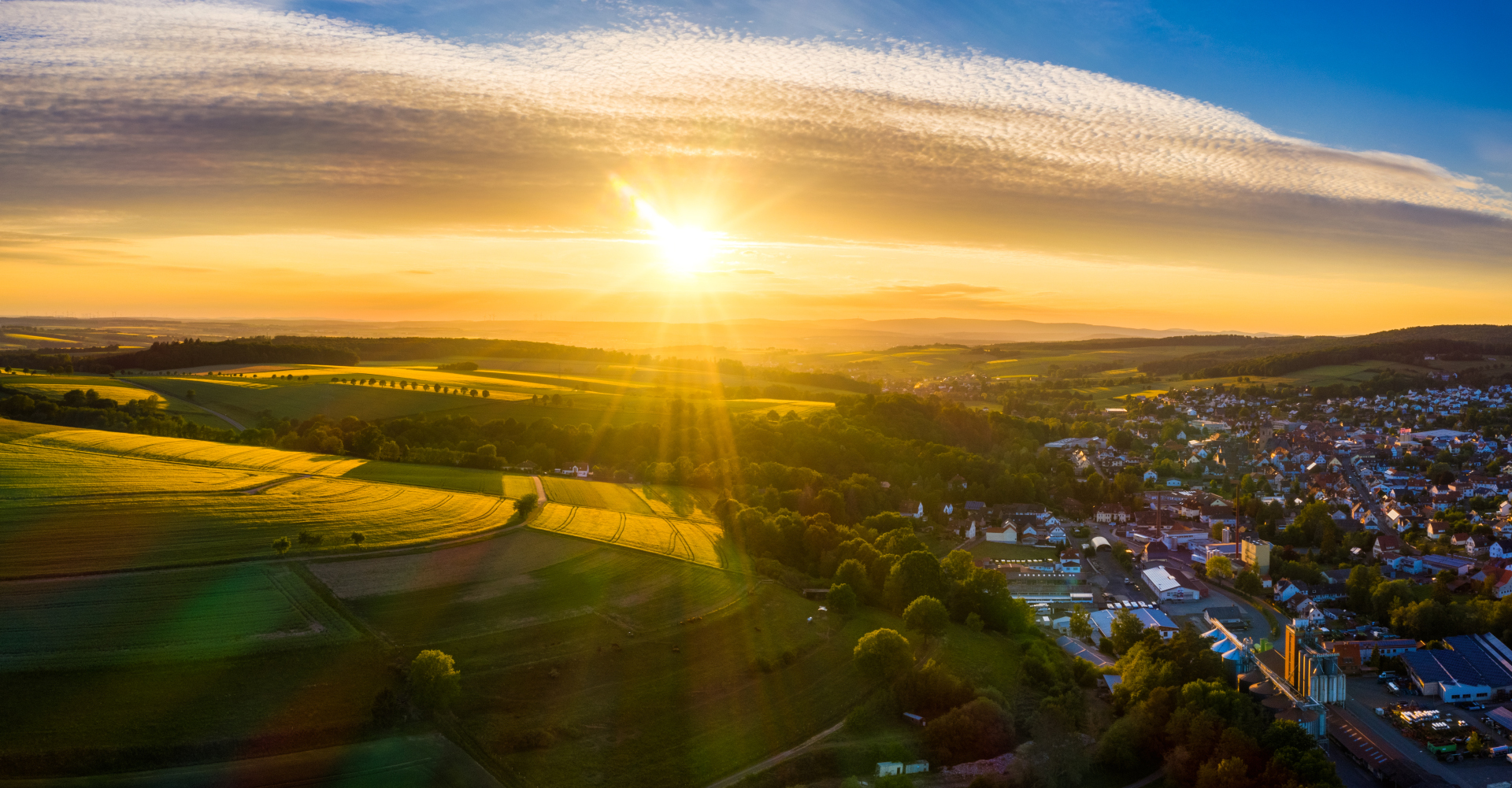 Sonnenuntergang über der Stadt Neukirchen; Im linken unteren Bereich ist eine hügelige Graslandschaft zu sehen, rechts daneben Wohnhäuser der Stadt Neukirchen. Im oberen Teil ist der Himmel mit Sonnenuntergang zu sehen.