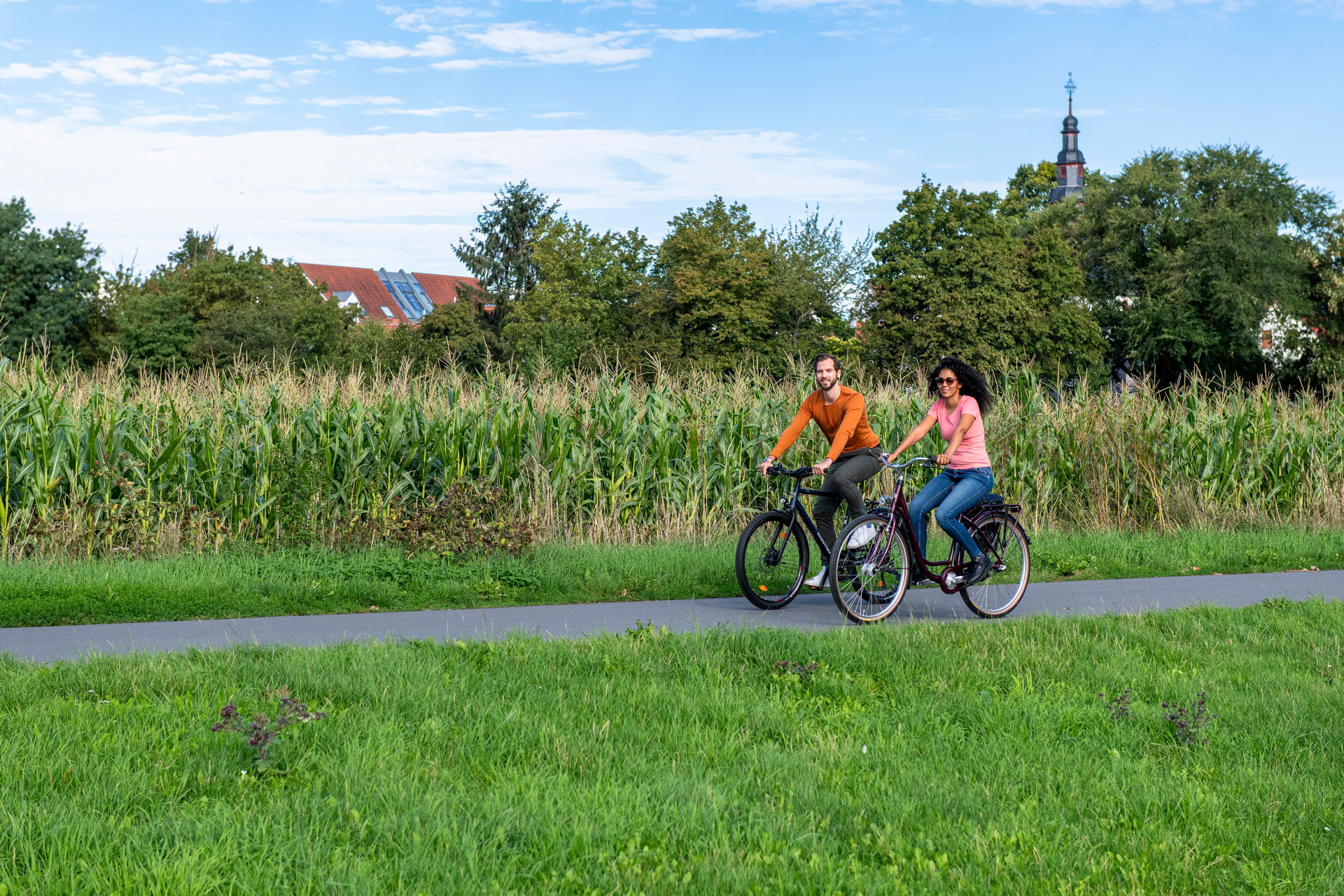 Zwei radfahrende Personen auf einem Fahrradweg. Im Vordergrund eine Wiese, im Hintergrund ein Maisfeld, dahinter ein Kirchturm sowie das Dach eines Gebäudes.