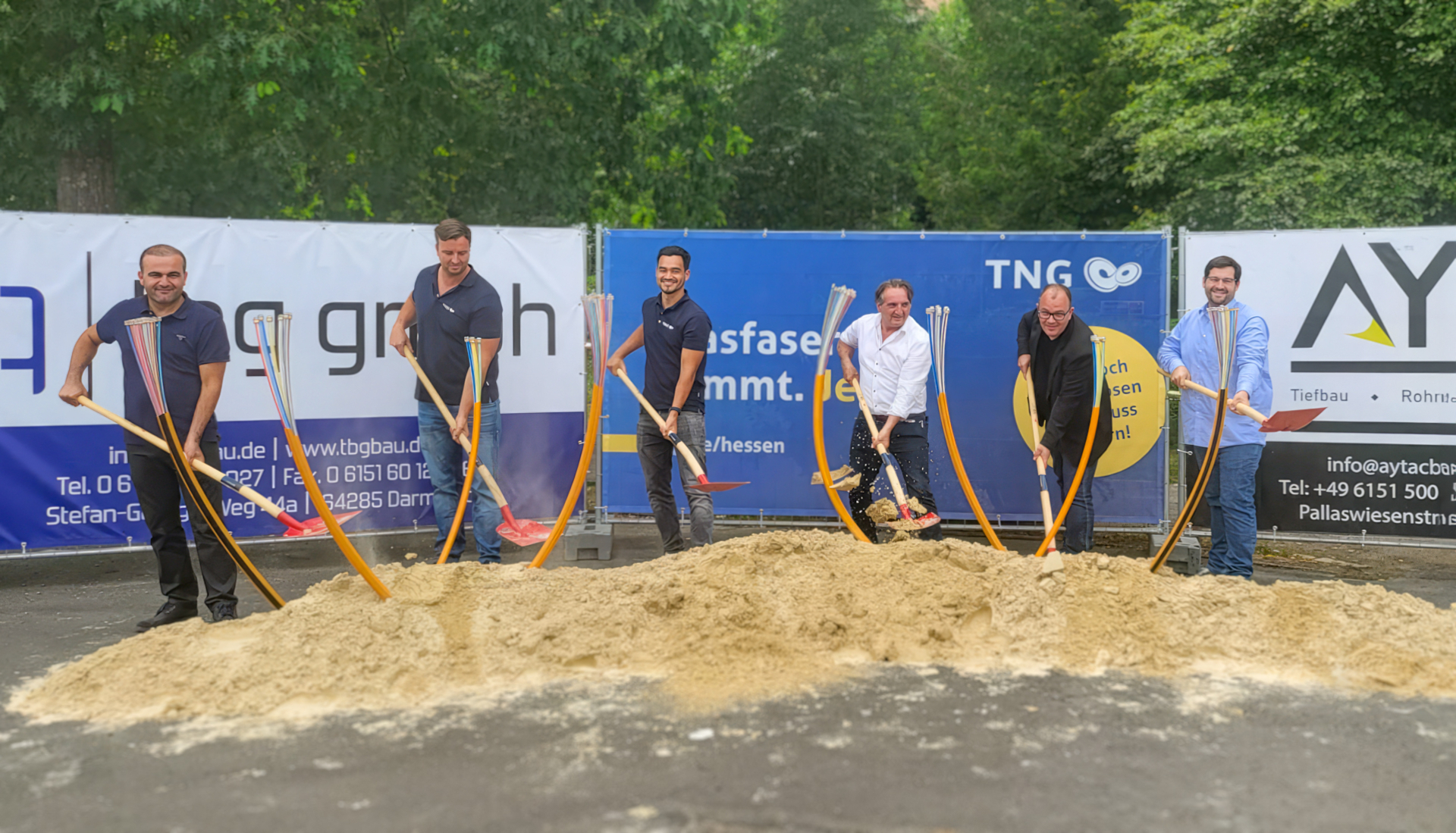 Foto (v. l. n. r.): Bekir Turan (Geschäftsführer TBG GmbH), Raphael Kupfermann (TNG-Regionalleiter), Juan Torp (TNG-Netzplaner), Sükrü Tasdemir (Projektleiter Aytac Bau GmbH), Marian Knauff (Bürgermeister Neukirchen), Jörn Szypkowski (Digitalisierungsbeauftragter Neukirchen). stehen mit Spaten hinter einen Haufen Sand. Im Sand stecken kurze Stücke von Glasfaserleerrohren.