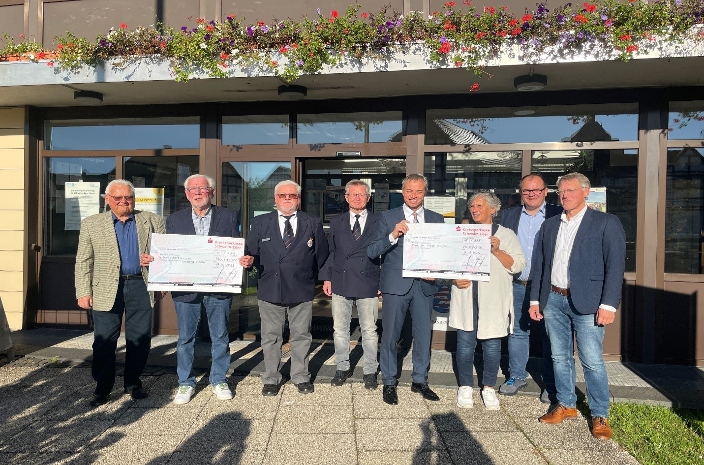 v.l.Dieter Fischer, Dieter Kirchner, Wolfgang Hertel, Heinz Roß, Bürgermeister Michael Merz, Constanze Freund, Bürgermeister Marian Knauff, Joachim Freund stehen vor dem Eingang des Rathauses Neukirchen. Dieter Kirchner sowie Bürgermeister Michael Merz gemeinsam mit Constanze Freund halten zwei großformatige Schecks in der Hand.