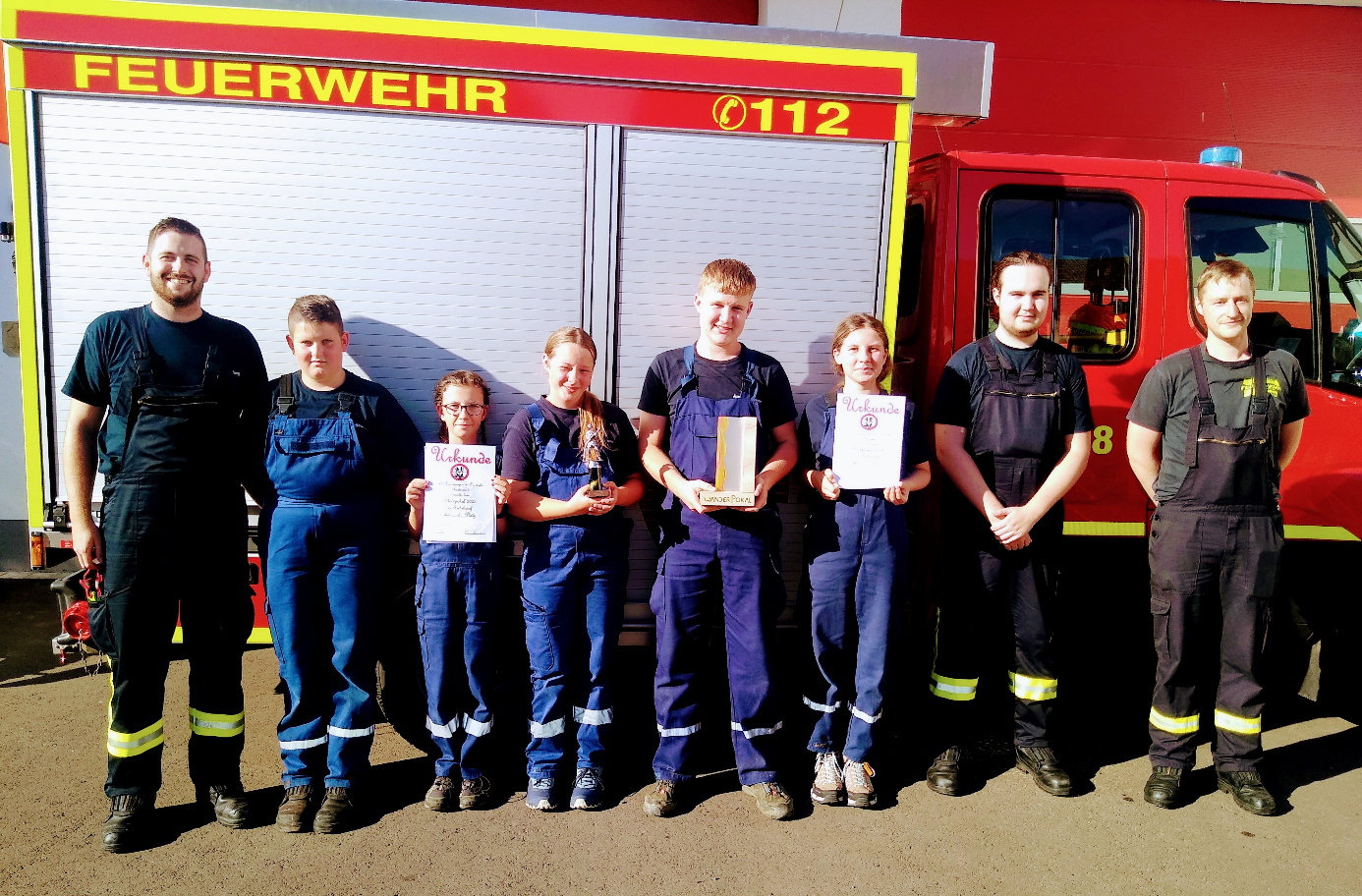 7 Jugendliche der Jugendfeuerwehr stehen mit Ihrem Betreuer vor einem Feuerwehrfahrzeug. Einige halten Urkunden in der Hand.