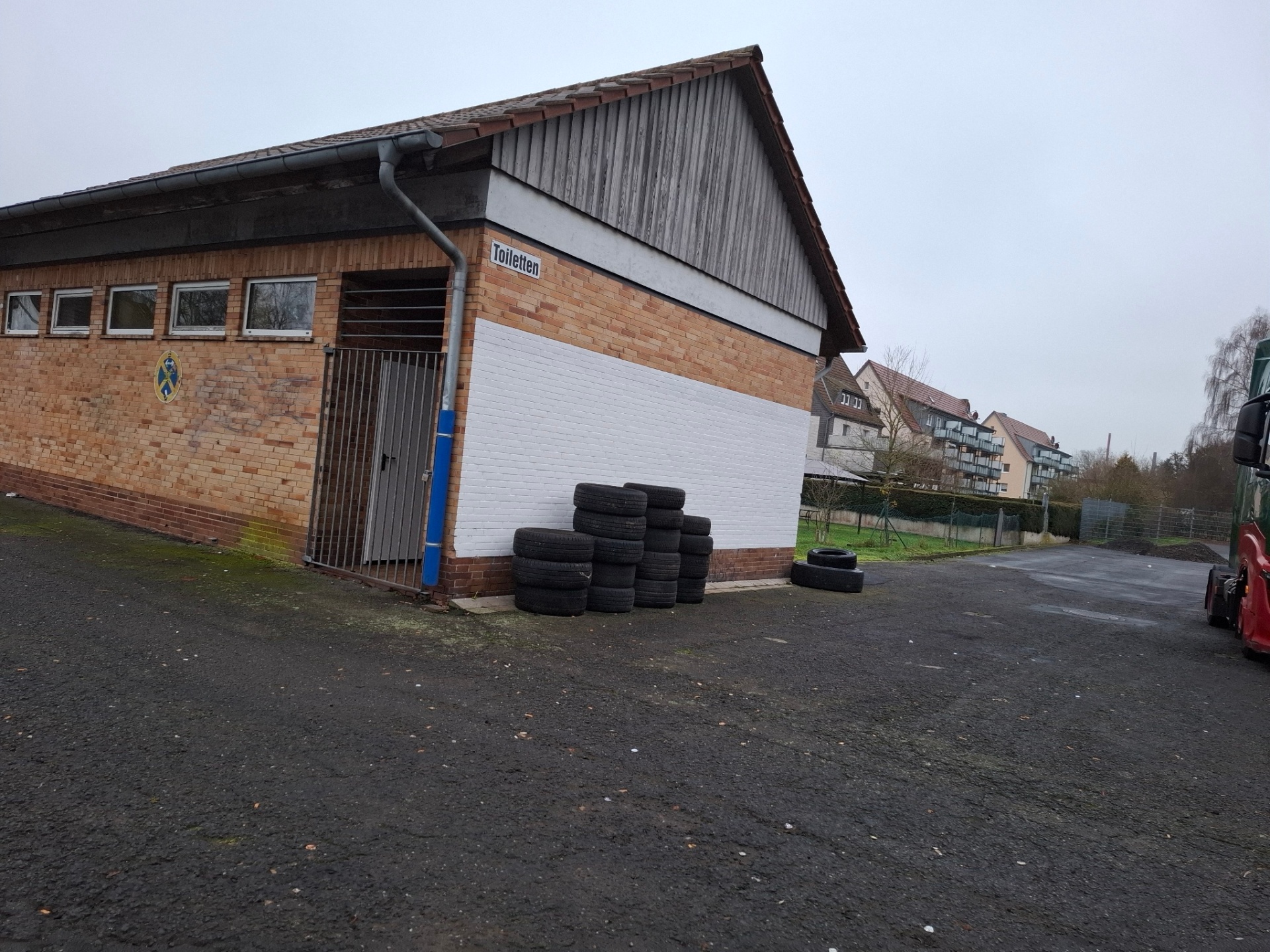 Seitliche Ansicht eines eingeschossigen Gebäudes mit Ziegelwänden und Satteldach. An der rechten Wand ist ein weiß gestrichener Bereich zu sehen, davor stehen mehrere gestapelte Autoreifen. Links befindet sich ein verschlossener Eingang mit Metallgitter, darüber ein kleines Schild mit der Aufschrift „Toiletten“. Das Gebäude steht auf einem asphaltierten Hof; im Hintergrund sind Wohnhäuser und Bäume erkennbar.