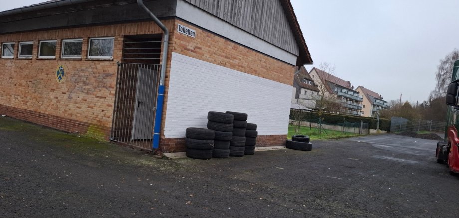 Seitliche Ansicht eines eingeschossigen Gebäudes mit Ziegelwänden und Satteldach. An der rechten Wand ist ein weiß gestrichener Bereich zu sehen, davor stehen mehrere gestapelte Autoreifen. Links befindet sich ein verschlossener Eingang mit Metallgitter, darüber ein kleines Schild mit der Aufschrift „Toiletten“. Das Gebäude steht auf einem asphaltierten Hof; im Hintergrund sind Wohnhäuser und Bäume erkennbar.