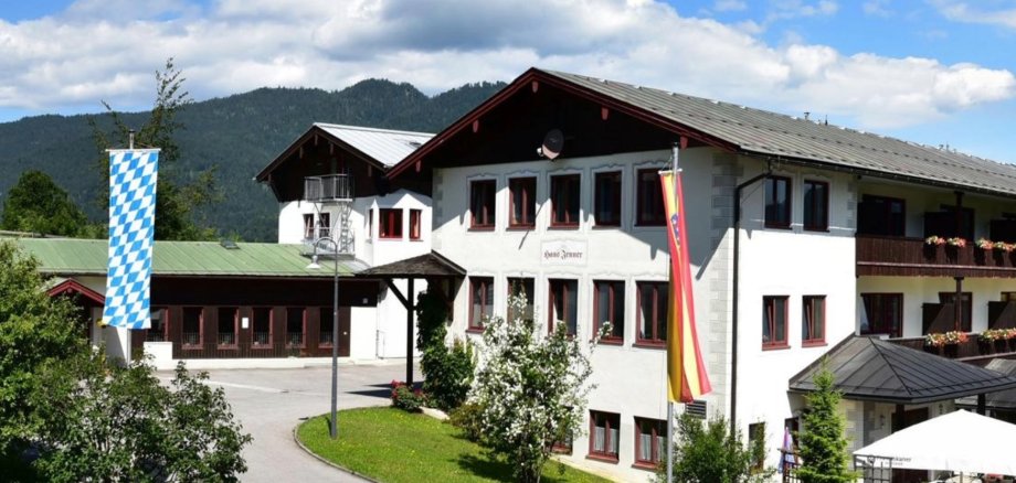 Im Vordergrund ist ein Gebäude mit der Aufschrift "Haus Jenner". Im Hintergrund ist ein weiteres Gebäude zu sehen. Dahinter eine bewaldete Bergkette. Links vorne ist eine blau-weiße Fahne.