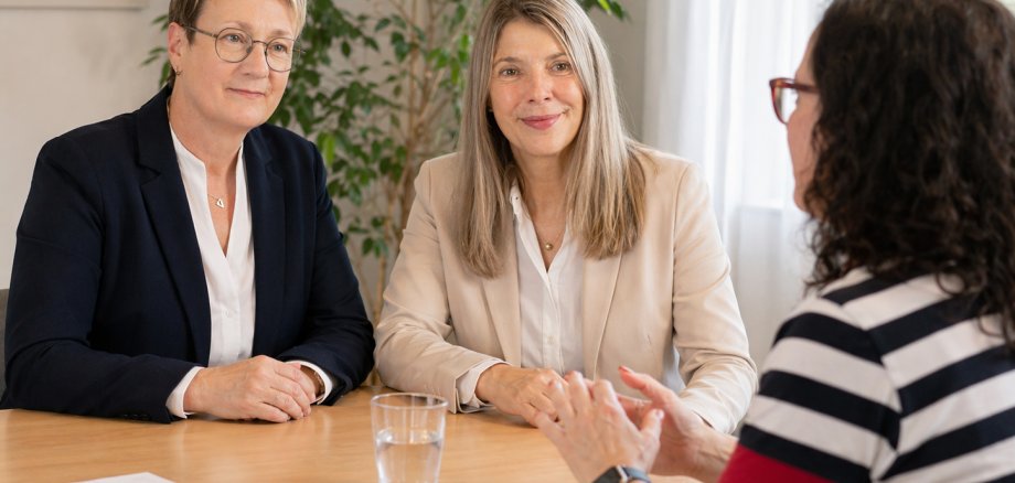 Auf dem Foto sind 3 Frauen zu sehen. Sie sitzen an einem Tisch. Zwei der Frauen hören der dritten zu.