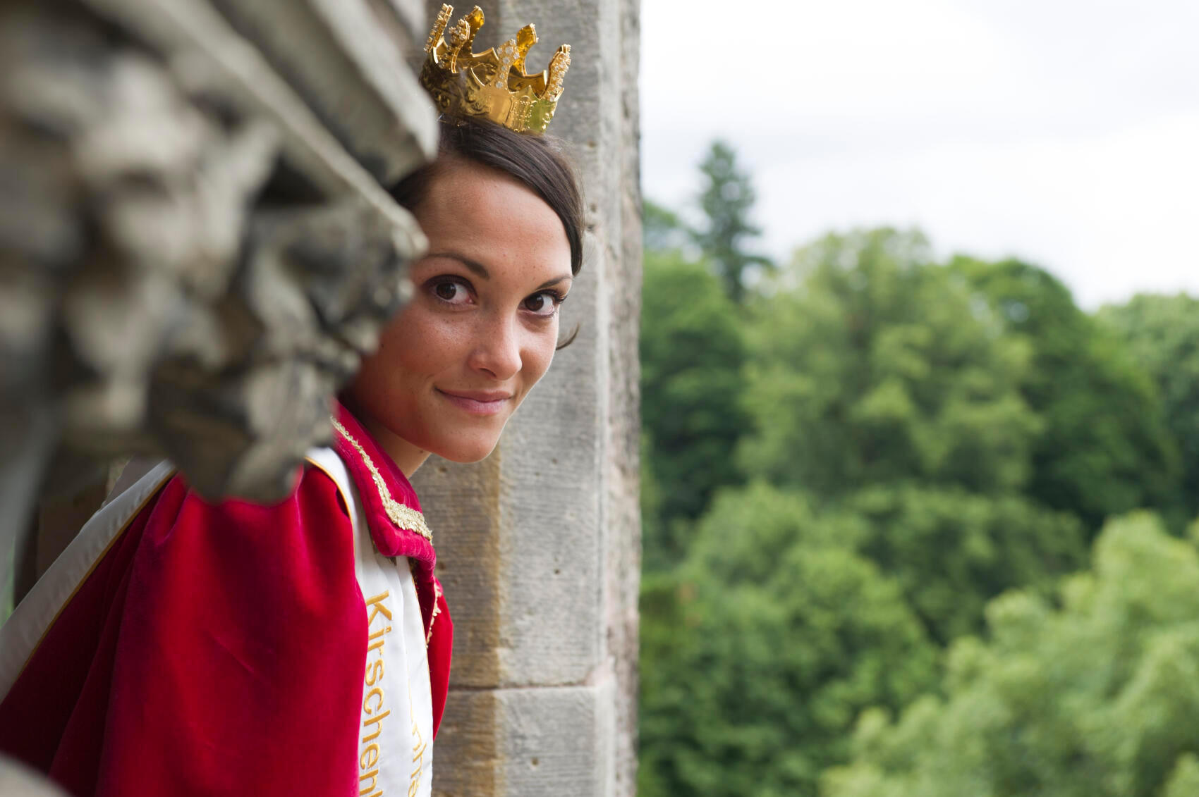 Auf dem Foto ist die Kirschenkönigin von Witzenhausen zu sehen - eine junge Dame mit dunklen Haaren, goldener Krone und einem roten Gewand. Sie schaut aus dem Fenster einer Burg.