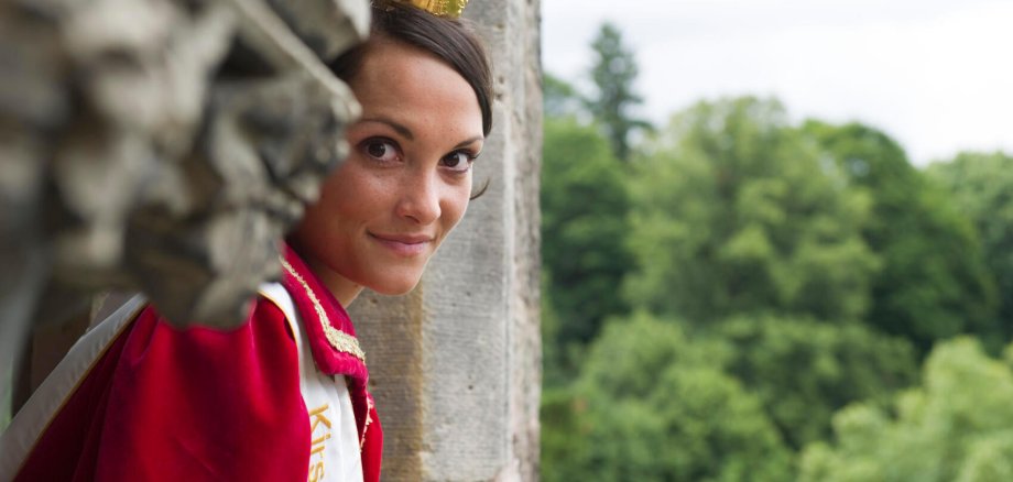 Auf dem Foto ist die Kirschenkönigin von Witzenhausen zu sehen - eine junge Dame mit dunklen Haaren, goldener Krone und einem roten Gewand. Sie schaut aus dem Fenster einer Burg.