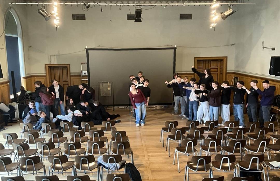 Auf dem Bild sind Jugendliche zu sehen, die für eine Theateraufführung proben. Im unteren Bereich des Bildes sind leere Stühle zu sehen.