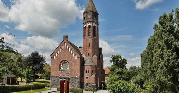 Auf dem Foto ist die Hephata-Kirche in Treysa zu sehen. Es ist eine rote Backsteinkirche mit hohem Kirchturm. Drum herum stehen einige grüne Baume.