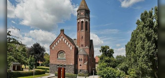 Auf dem Foto ist die Hephata-Kirche in Treysa zu sehen. Es ist eine rote Backsteinkirche mit hohem Kirchturm. Drum herum stehen einige grüne Baume.