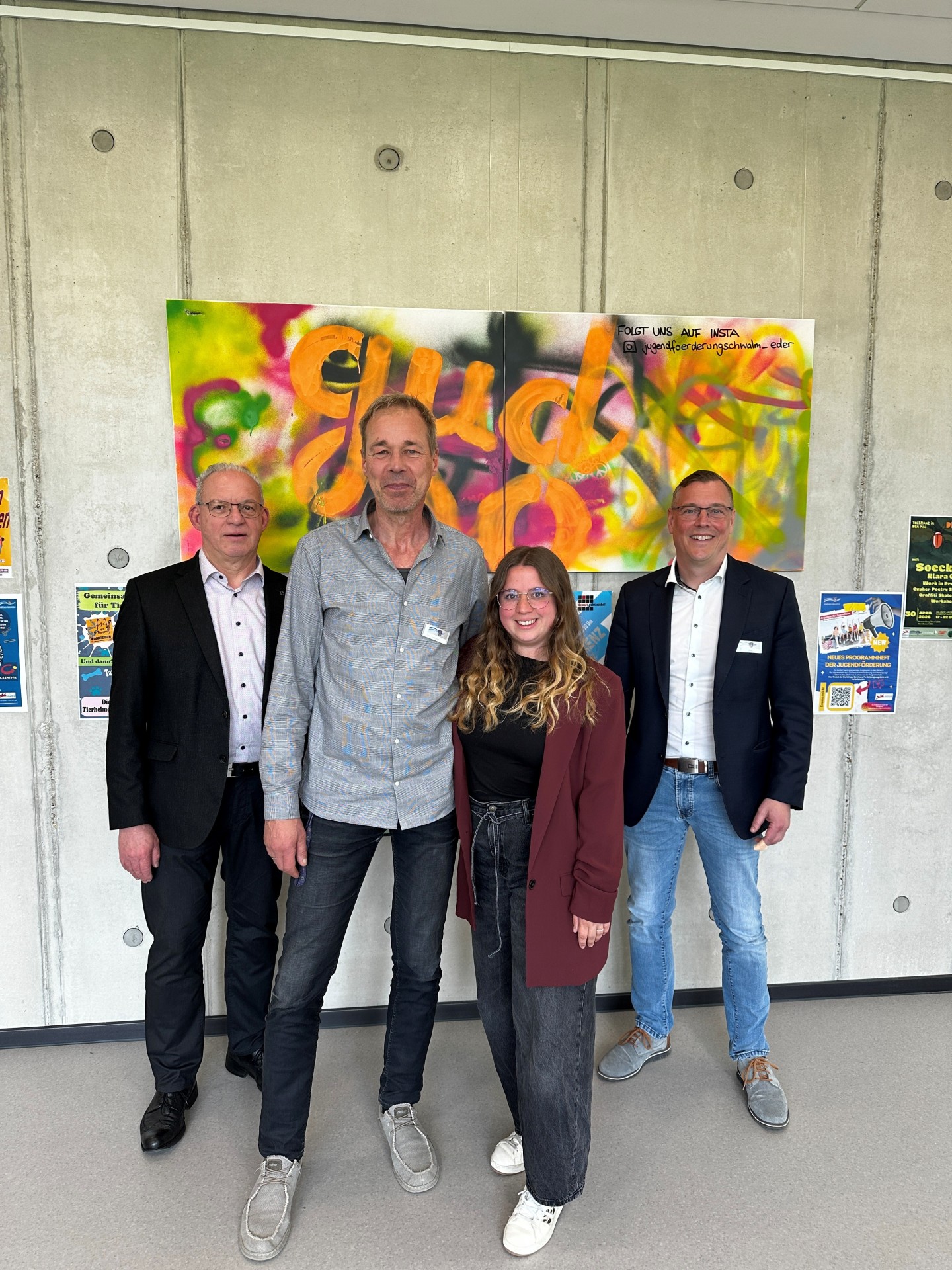Auf dem Foto sind drei Männer und eine Frau zu sehen. Im Hintergrund eine Botenwand mit einem Graffiti Bild.