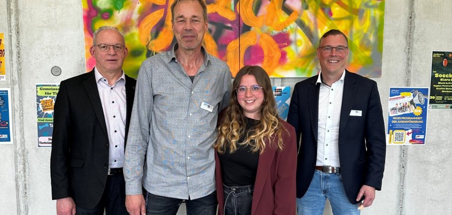 Auf dem Foto sind drei Männer und eine Frau zu sehen. Im Hintergrund eine Botenwand mit einem Graffiti Bild.