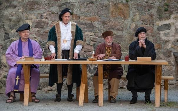 Auf dem Foto sind vier Männer in Verkleidung zu sehen. Drei sitzen an einem Tisch, einer steht. Es soll eine Theatergruppe darstellen, die ein Stück aus der Geschichte der Reformation spielt