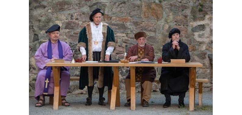 Auf dem Foto sind vier Männer in Verkleidung zu sehen. Drei sitzen an einem Tisch, einer steht. Es soll eine Theatergruppe darstellen, die ein Stück aus der Geschichte der Reformation spielt