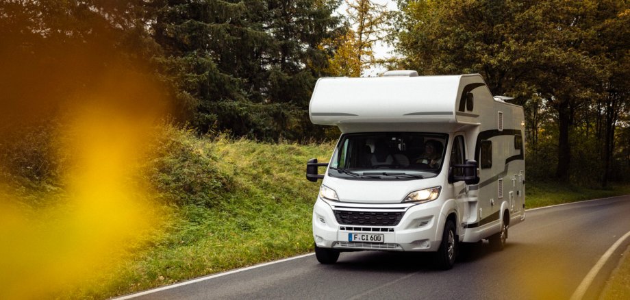Auf dem Foto ist ein Wohnmobil zu sehen, das auf einer Straße entlang fährt. Drum herum Bäume und grüne Wiese